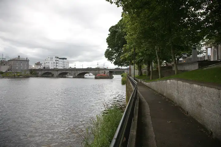 The stone arches of Athlone Town Bridge across the wide Shannon under a grey sky, with town buildings on the far bank and moored boats visible through the arches.