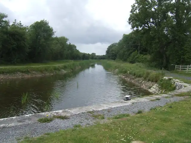 Een recht stuk kanaal bij Island Bridge loopt tussen met bomen omzoomde oevers, gezien vanaf een met steen omrande kade met één meerpaal.
