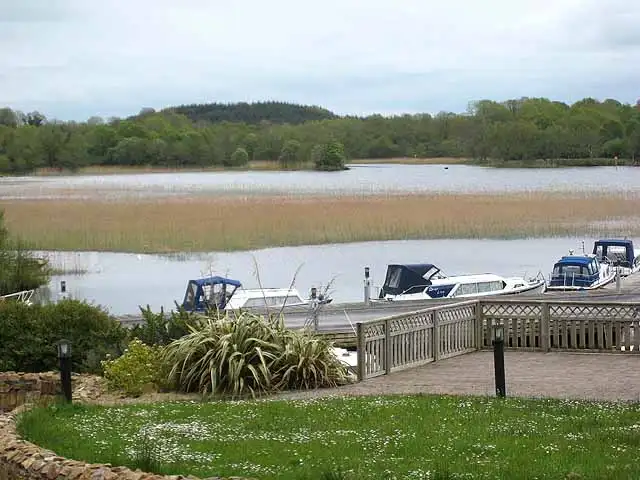 Motorboote liegen an einem Steg unterhalb einer Gartenterrasse am See, dahinter Schilfgürtel und das bewaldete Ufer des Lough Key.