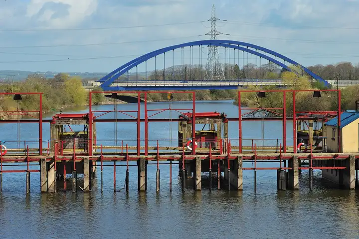 The red-painted sluice gates across the River Bann at Toome, with the blue arch of the bypass bridge and an electricity pylon behind them.