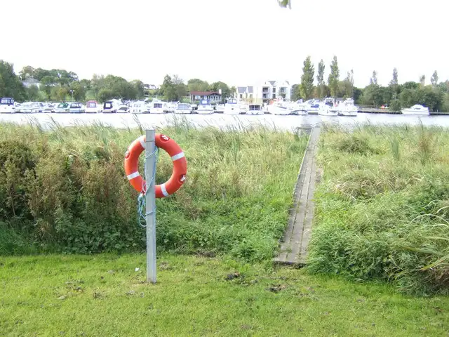 Rows of moored cruisers line the Bellanaleck marina across the River Erne, seen past a lifebuoy and a boardwalk through the reeds on the opposite bank.