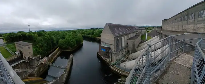 Depuis une passerelle en hauteur à Ardnacrusha, la vue plonge sur le canal de fuite, entre des murs de béton à gauche et la centrale avec ses conduites forcées à droite.