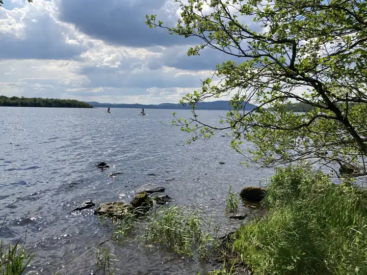 Sunlight glitters on the lough off a stony, grassy bank at Castle Archdale, with two small craft far out on the water and blue hills on the horizon.