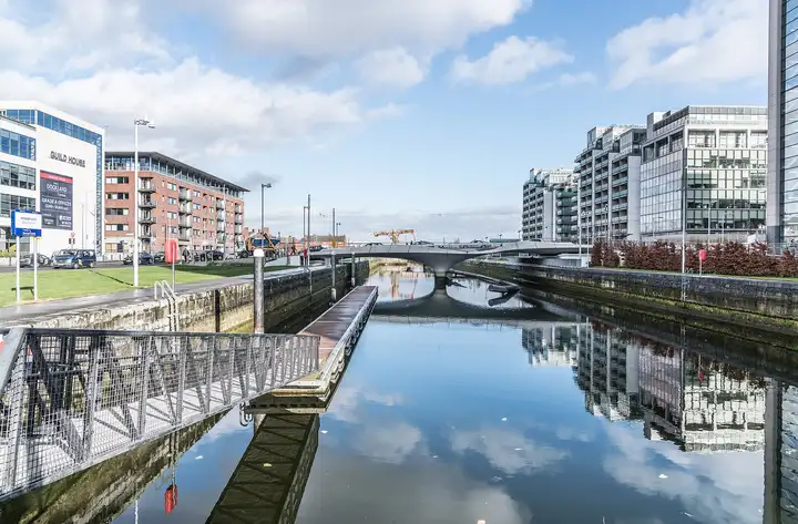 The still water of Spencer Dock mirrors blue sky, clouds and modern office and apartment blocks, with a floating pontoon on the left and the curved Spencer Dock Bridge across the dock ahead.