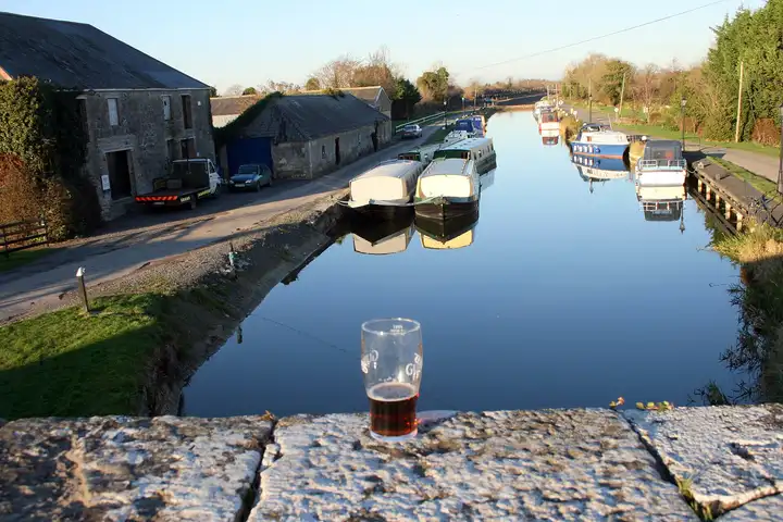 Boats moored along both banks of the Barrow Line at Vicarstown, seen from the bridge, with a pint glass on the parapet and old stone stores beside the canal.