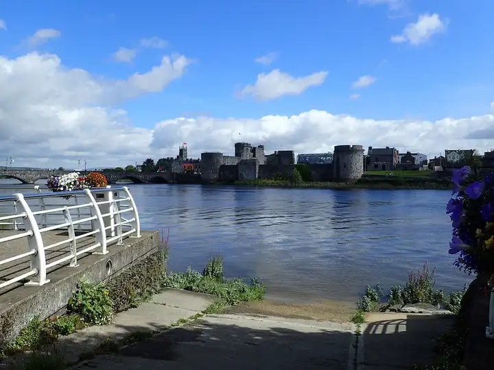 King John’s Castle liegt jenseits des breiten Shannon, gesehen von einer Slipanlage am Ufer mit weißem Geländer und Blumenkörben.