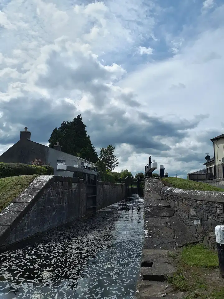 Die steinerne Kammer der 16. Schleuse in Kilcock zieht sich bis zu den oberen Toren, links ein graues Häuschen und dunkle Bäume unter einem weiten, bewölkten Himmel.